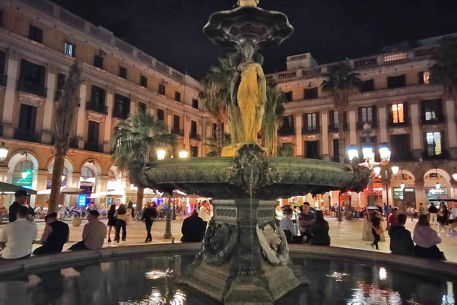 Der Drei-Grazien-Brunnen in der Mitte der Plaça Reial bei Nacht, von unten beleuchtet, mit Arkadengebäuden im Hintergrund