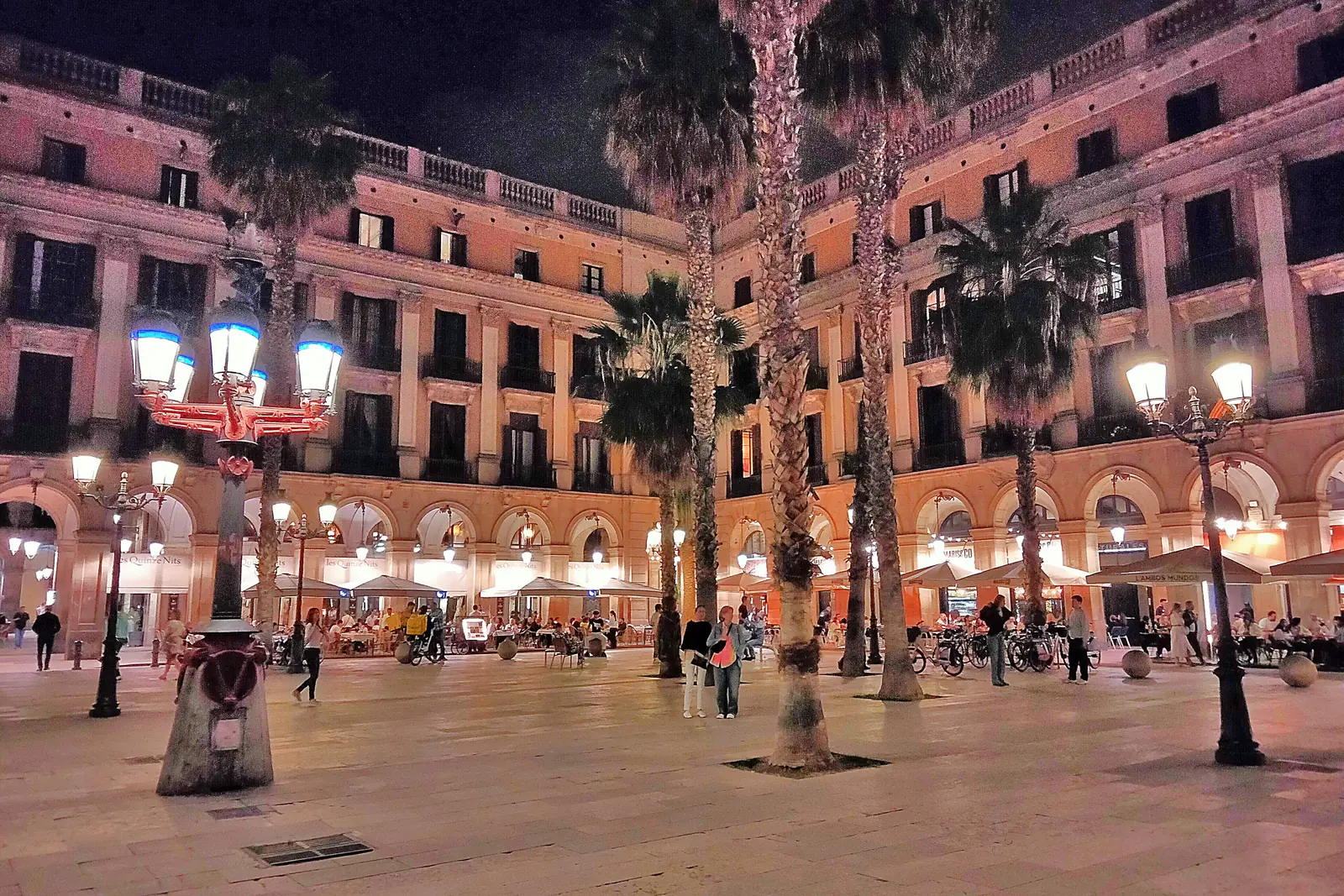 Eine der Straßenlaternen, die Antoni Gaudí für die Plaça Reial entworfen hat, bei Nacht beleuchtet, mit Palmen und Arkadengebäuden im Hintergrund