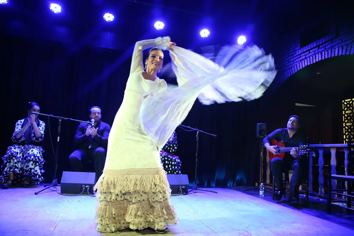 Tanečnice flamenca v dlouhých bílých třásnitých šatech rozvíří šál na osvětleném pódiu, zatímco ji doprovází zpěvák a kytarista