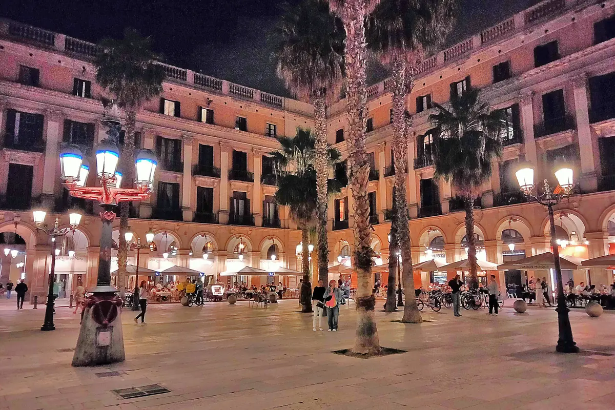 Jedna z pouličních lamp, které Antoni Gaudí navrhl pro Plaça Reial, rozsvícená v noci, v pozadí palmy a budovy s arkádami