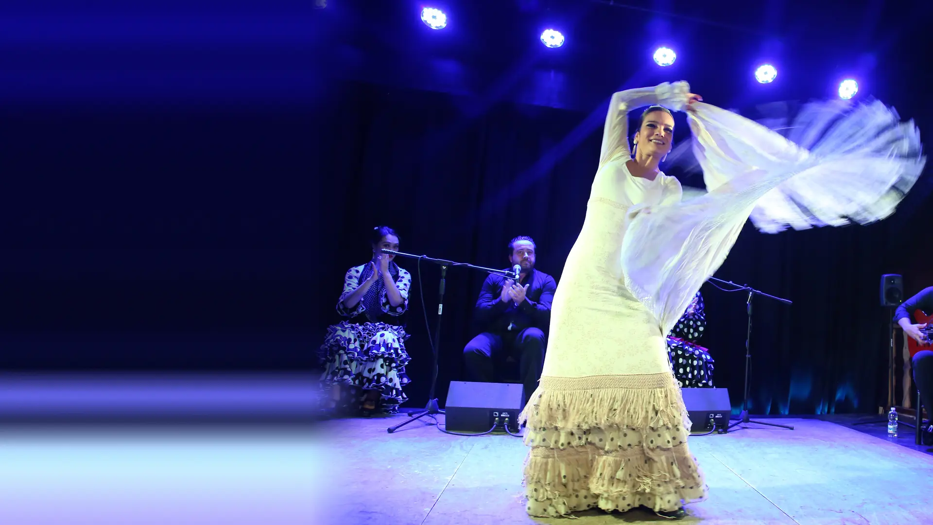 Eine Flamenco-Tänzerin in einem langen weißen Fransenkleid lässt einen hellen Schal über eine blau beleuchtete Bühne gleiten, während hinter ihr ein Sänger und ein Gitarrist spielen