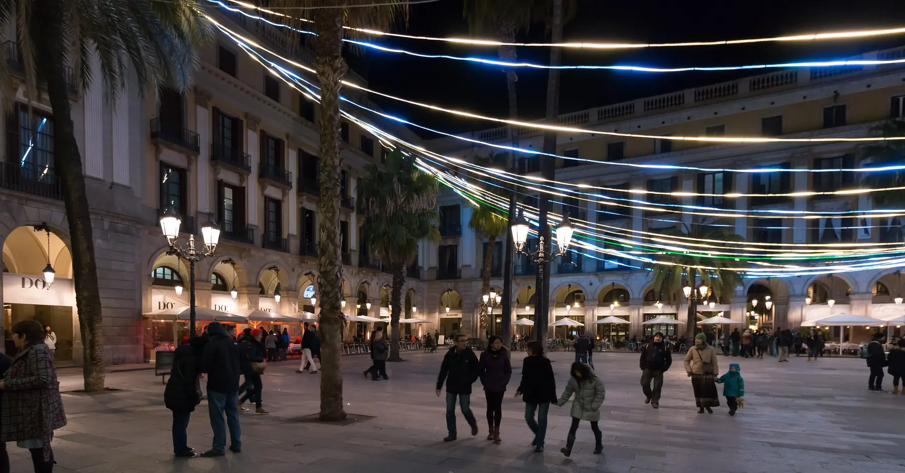 Plaça Reial bei Nacht, mit Lichterketten, die zwischen Palmen über einem Platz mit Straßenlaternen und Arkadengebäuden gespannt sind